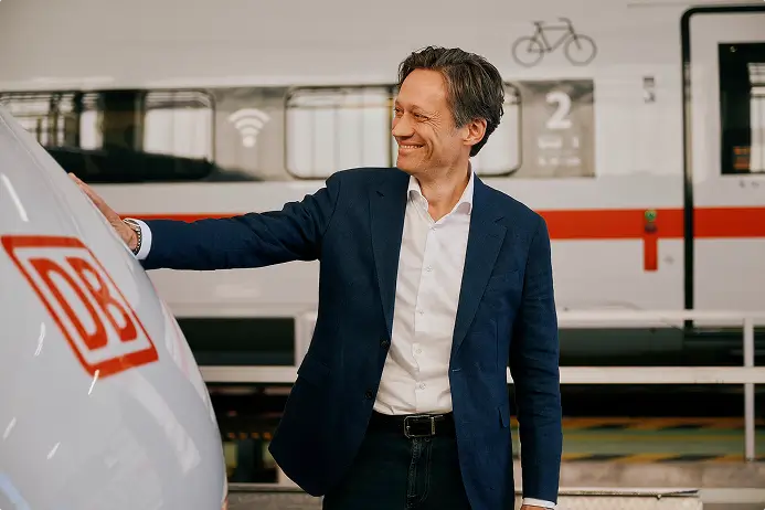 Eine Person im blauen Sakko lehnt an einem ICE-Zug, im Hintergrund ist ein weiterer Zug zu sehen.