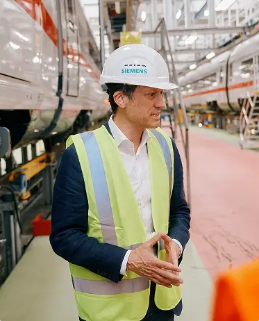 Eine Person mit Schutzweste und Helm steht vor einem ICE-Zug im Werk und spricht mit anderen.