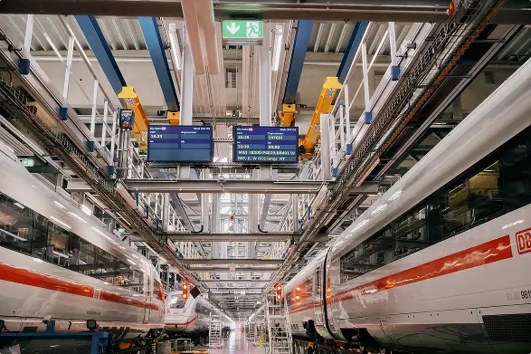 Zwei ICE-Züge stehen sich in einer großen, modernen Werkhalle mit Anzeigen und Technik gegenüber.