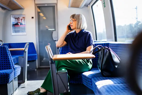 Eine Frau mit weißem Haar telefoniert und sitzt mit einer schwarzen Tasche im Zugabteil.