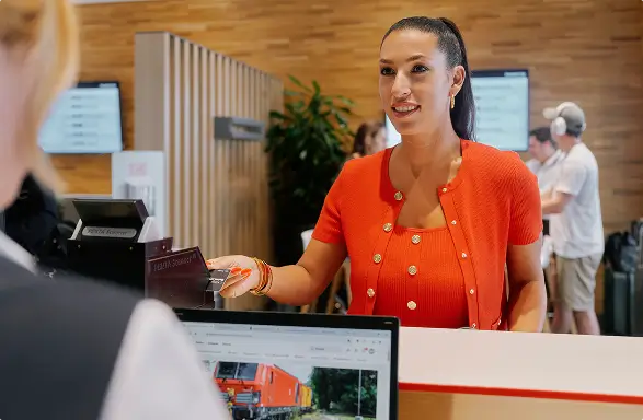 Frau im roten Kleid am Empfang einer DB Lounge, im Hintergrund weitere Reisende.