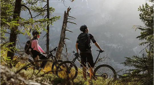 Zwei Mountainbiker stehen mit ihren Fahrrädern am Waldrand und blicken auf eine Berglandschaft.