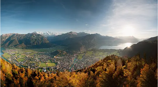 Panoramaaufnahme von Interlaken mit See, Stadt, herbstlichen Bäumen und umliegenden Bergen.