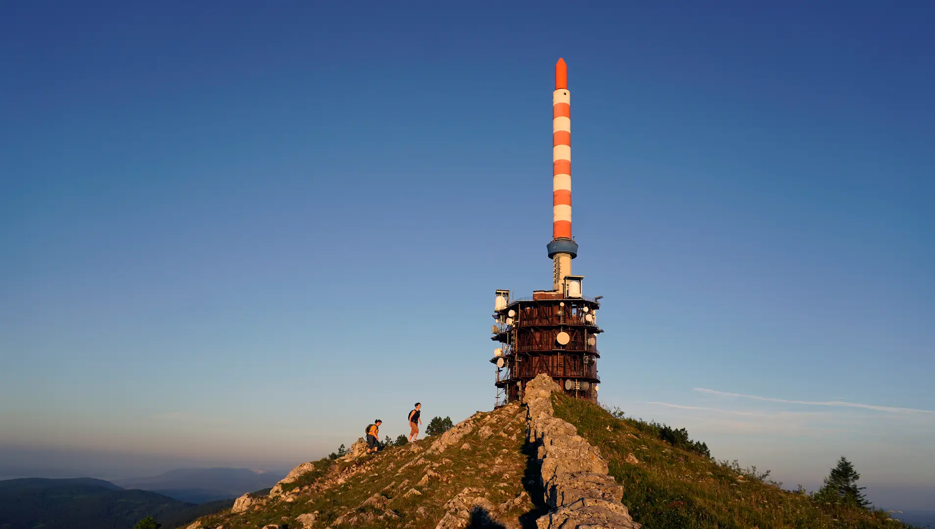 Großer Sendeturm auf einem Berggipfel mit Wanderern im Vordergrund und klarem Himmel.