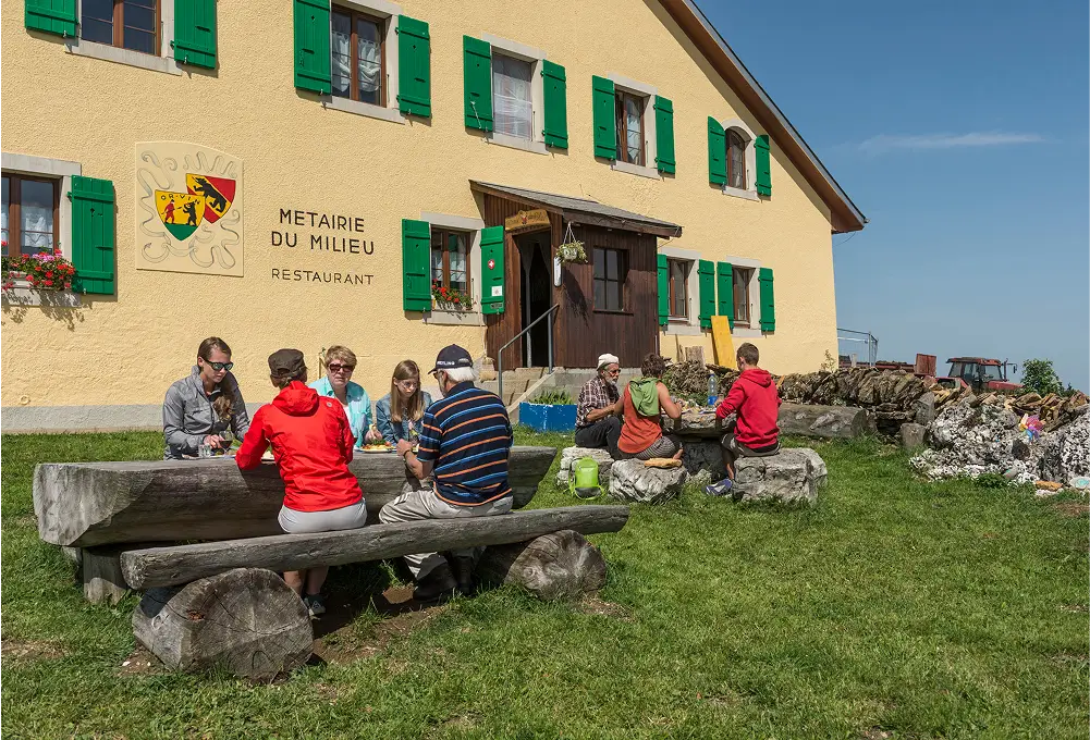 Gruppe von Menschen sitzt an Holztischen vor einem gelben Landgasthof mit grünen Fensterläden.