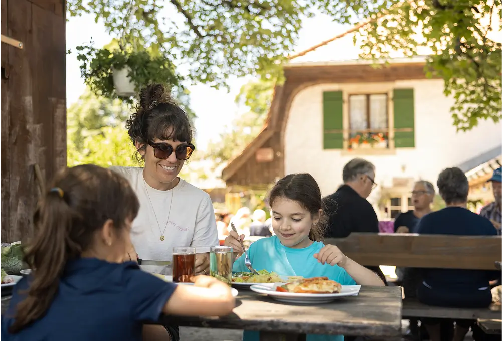 Eine Familie sitzt draußen an einem Holztisch und genießt gemeinsam ein Essen im Garten eines Landgasthofs. Im Hintergrund ist ein traditionelles Haus mit grünen Fensterläden zu sehen.
