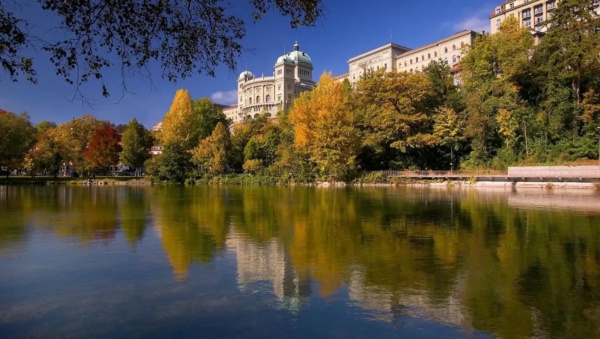 Das Bundeshaus in Bern spiegelt sich im ruhigen Wasser der Aare, umgeben von herbstlich gefärbten Bäumen.