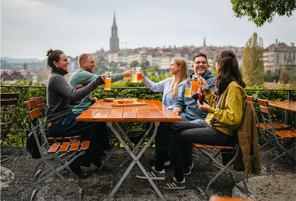 Eine Gruppe von Menschen stößt mit Biergläsern an einem Holztisch im Freien an, im Hintergrund ist die Altstadt von Bern mit dem markanten Münsterturm zu sehen.