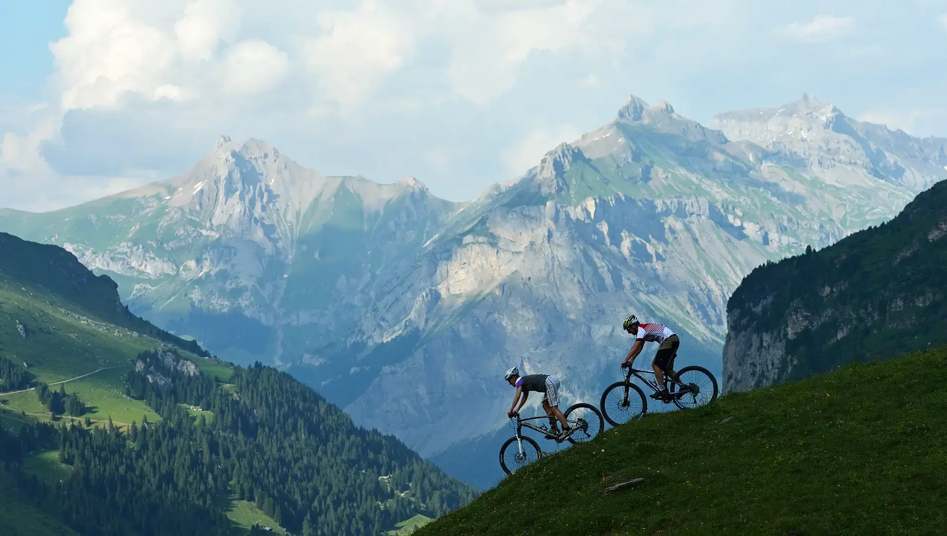 Zwei Mountainbiker fahren auf einem grasbewachsenen Berghang, umgeben von einer imposanten Alpenszenerie.