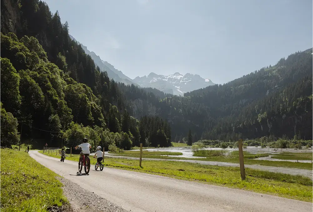 Kinder fahren auf einer ruhigen Straße durch ein grünes Tal mit Fluss, umgeben von bewaldeten Berghängen.
