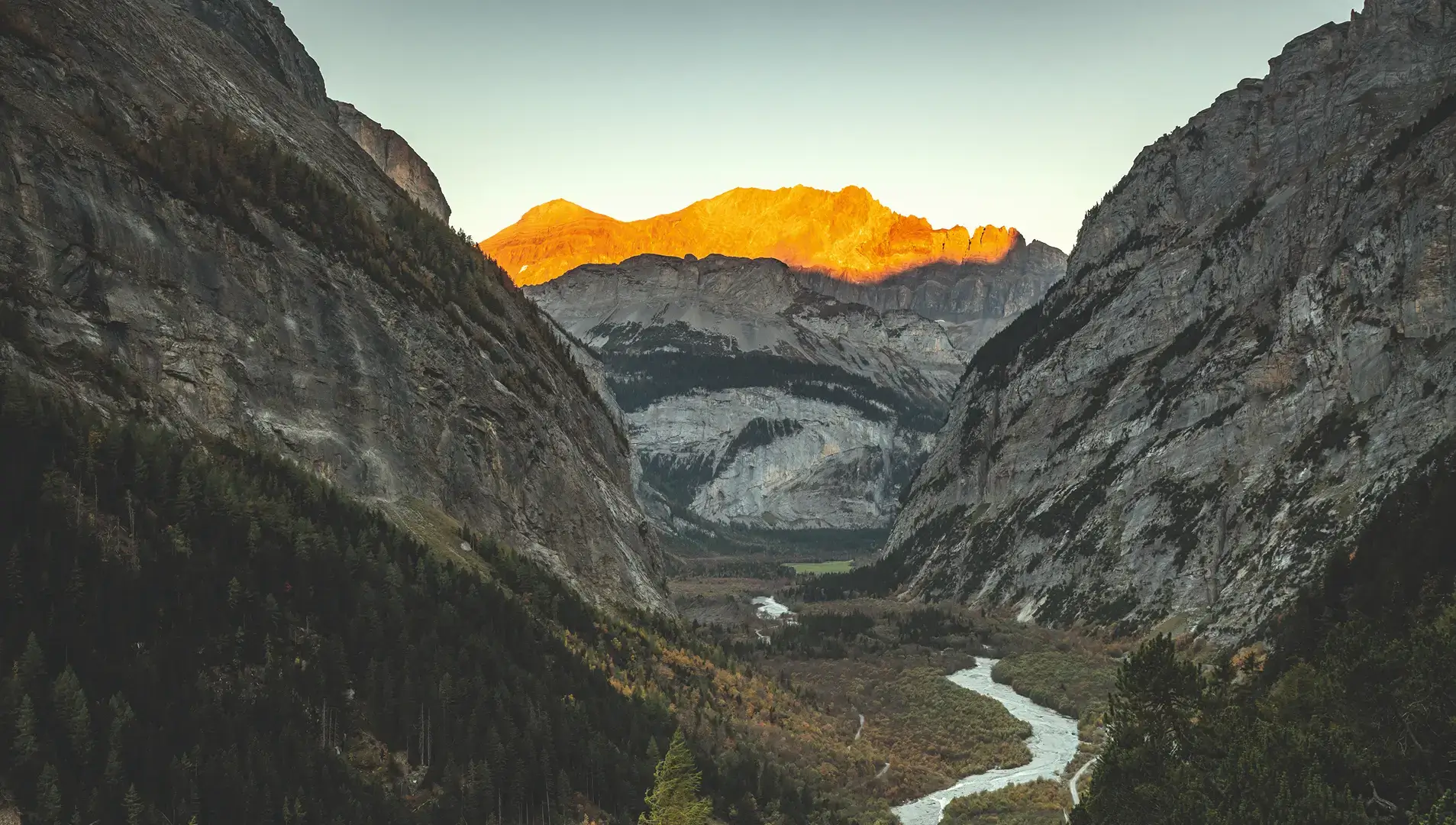 Sonnenaufgang über einem Flusstal zwischen steilen Felswänden, die Gipfel im Hintergrund sind in warmes Licht getaucht.