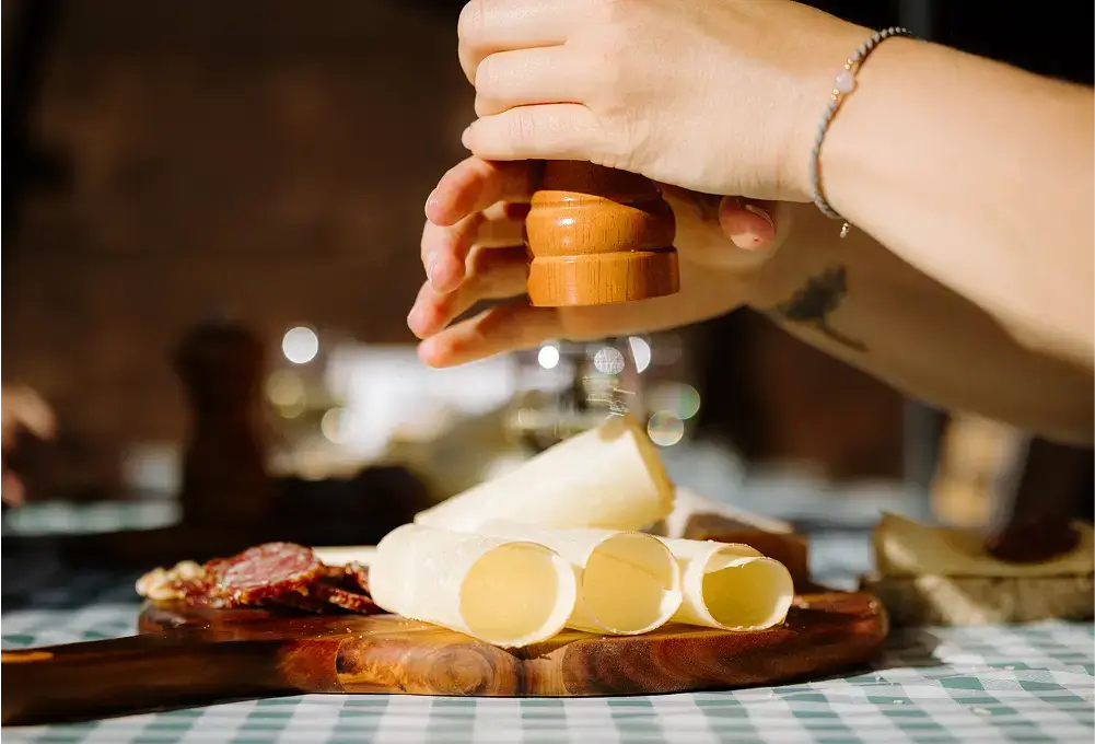 Auf einem Holzteller liegen eingerollte Käsescheiben und Wurst, eine Hand würzt das Essen mit einer Pfeffermühle.