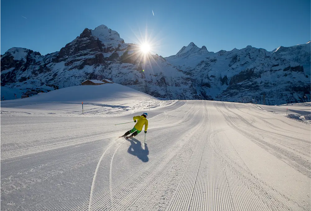 Skifahrer fährt auf einer frisch präparierten Piste vor beeindruckender Bergkulisse im Sonnenschein.