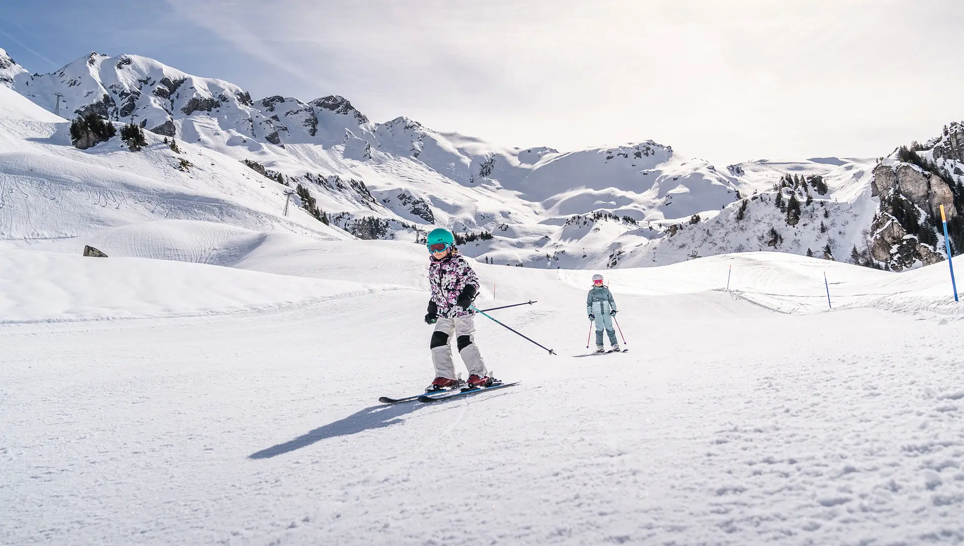 Zwei Kinder fahren auf Skiern eine breite, verschneite Piste inmitten einer alpinen Winterlandschaft hinab.