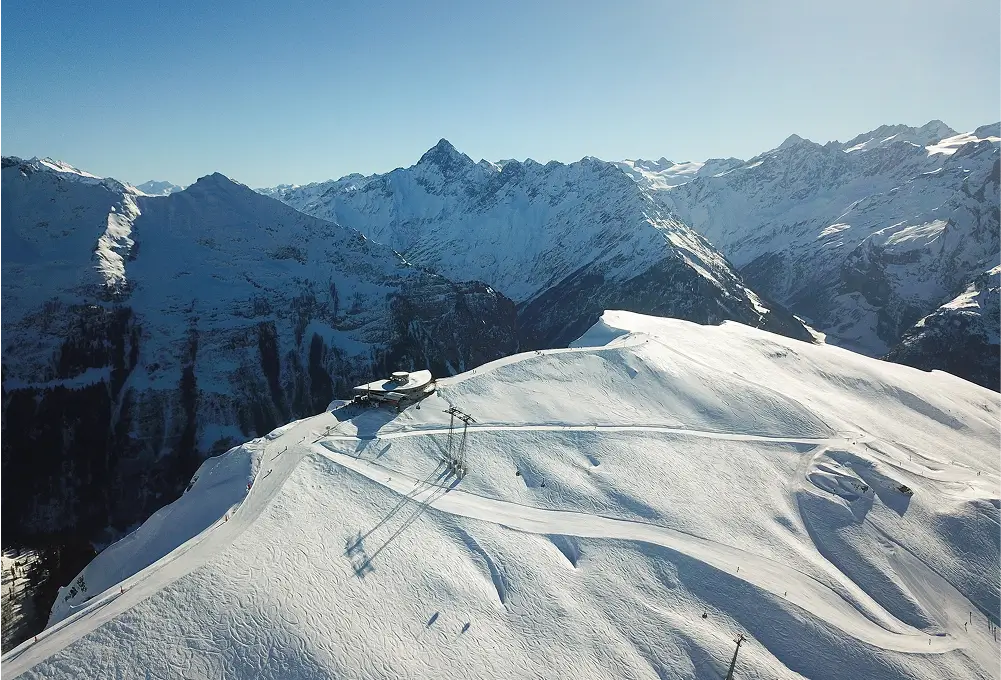 Eine verschneite Berglandschaft mit Skipisten und einer Bergstation, umgeben von hohen, schneebedeckten Alpen. Im Hintergrund erstreckt sich eine beeindruckende Gebirgskette unter klarem Himmel.