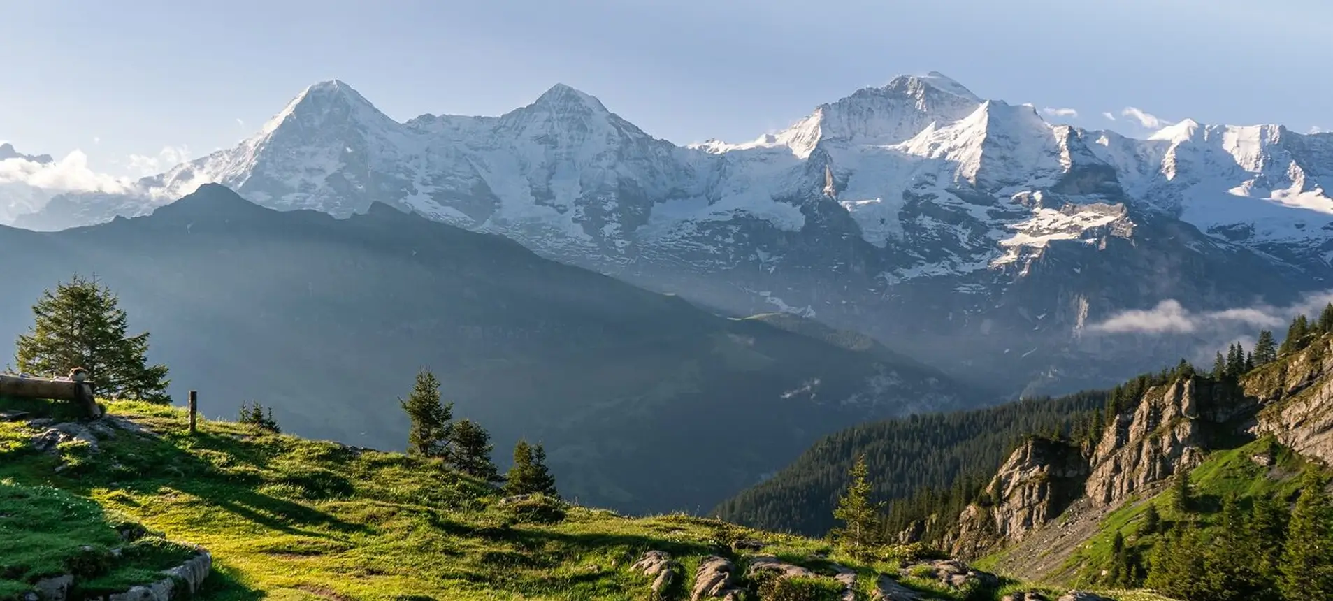 Zwei Wanderer spazieren bei Sonnenuntergang durch ein herbstliches Tal mit schneebedeckten Bergen im Hintergrund.