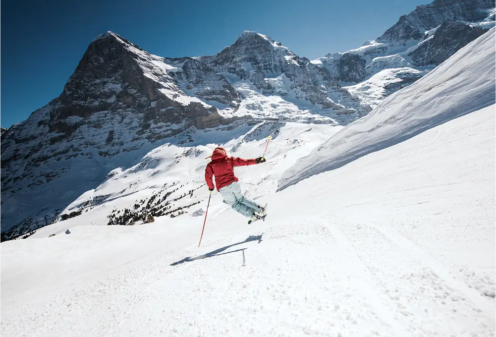 Ein Skifahrer in roter Jacke fährt auf einer verschneiten Piste vor majestätischer Bergkulisse.