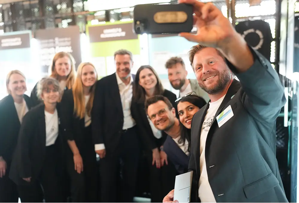 Eine Gruppe von Menschen posiert gemeinsam für ein Selfie bei einer Veranstaltung, im Hintergrund sind Infotafeln zu sehen.