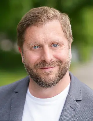 Ein Mann im Sakko steht in einem Innenraum und blickt in die Kamera, im Hintergrund sind Lichter und Veranstaltungselemente sichtbar.