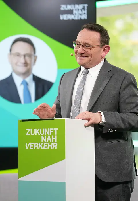 Ein Redner hält eine Präsentation an einem Pult mit der Aufschrift „Zukunft Nah Verkehr“ vor einer Leinwand mit grafischen Elementen.