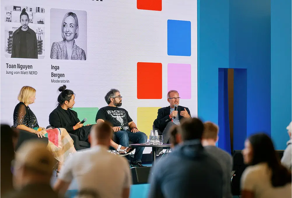 Podiumsdiskussion mit mehreren Personen auf einer Bühne, im Hintergrund ein großes Display mit Namen und Fotos der Teilnehmenden.
