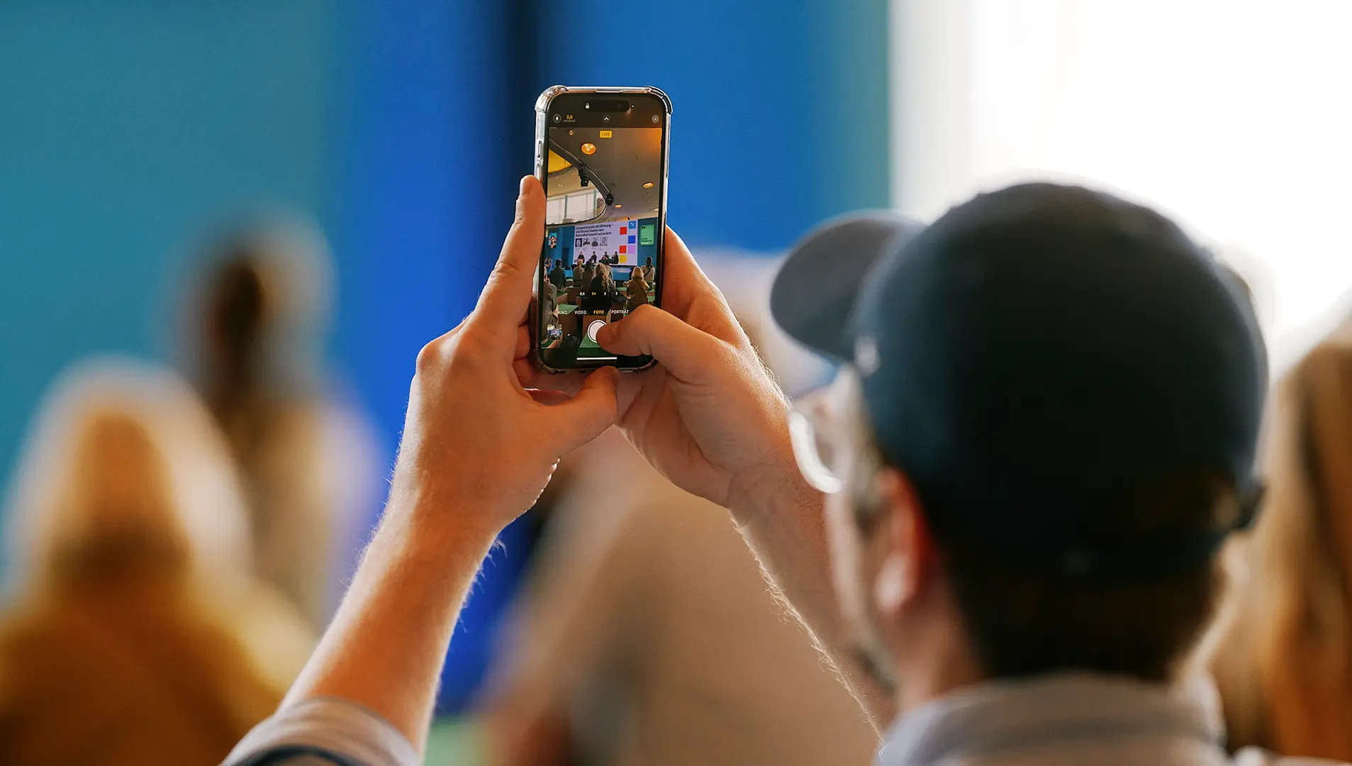 Eine Person fotografiert mit dem Smartphone eine Veranstaltung oder Podiumsdiskussion, die im Hintergrund stattfindet.