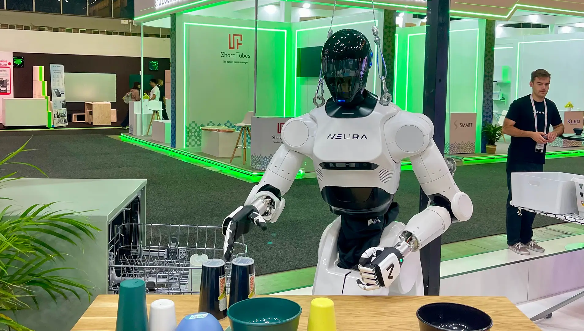 Ein humanoider Roboter der Marke Neura steht auf einer Messe und sortiert Geschirr auf einem Tisch. Im Hintergrund sind beleuchtete Messestände zu sehen.