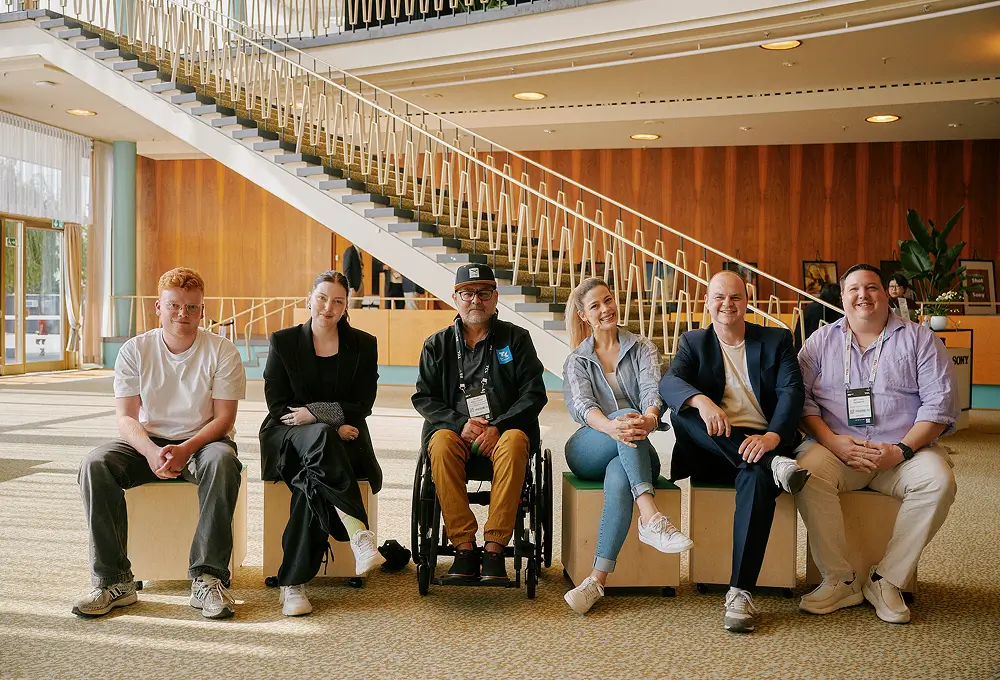 Sechs Personen sitzen nebeneinander in einer modernen Empfangshalle mit Treppe im Hintergrund.