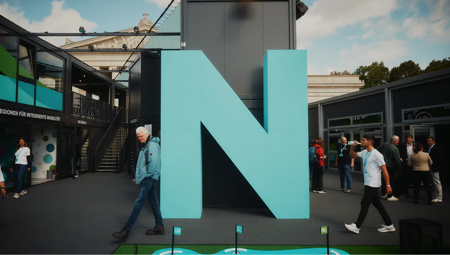 Eine große, türkisfarbene Skulptur des Buchstabens „N“ steht auf einem Messegelände, umgeben von Besuchern.