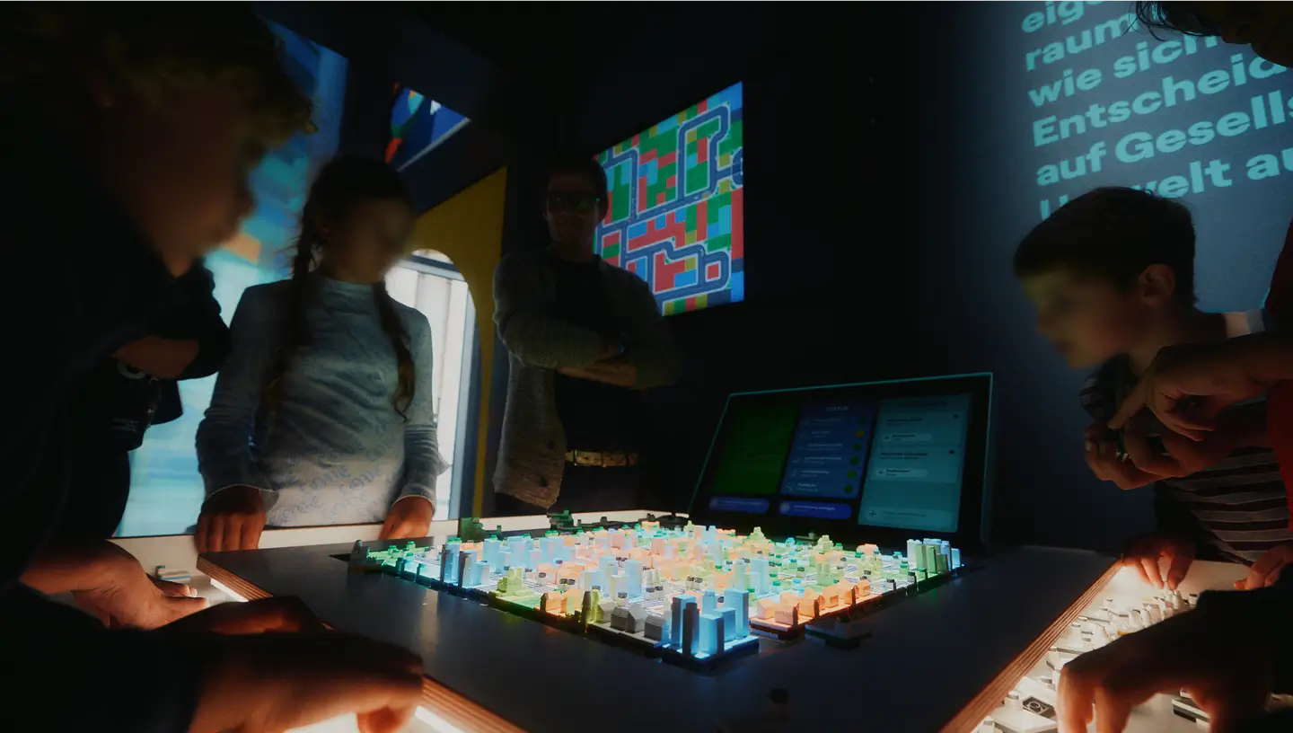 Mehrere Kinder stehen um ein interaktives, beleuchtetes Stadtmodell und bedienen gemeinsam eine digitale Ausstellungstafel.