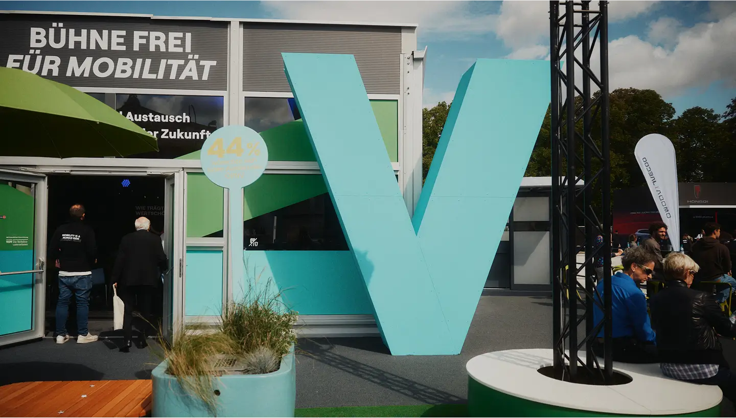 Eine große, türkisfarbene „V“-Skulptur steht vor einem Gebäude, Menschen betreten das Gebäude und sitzen auf Stühlen.