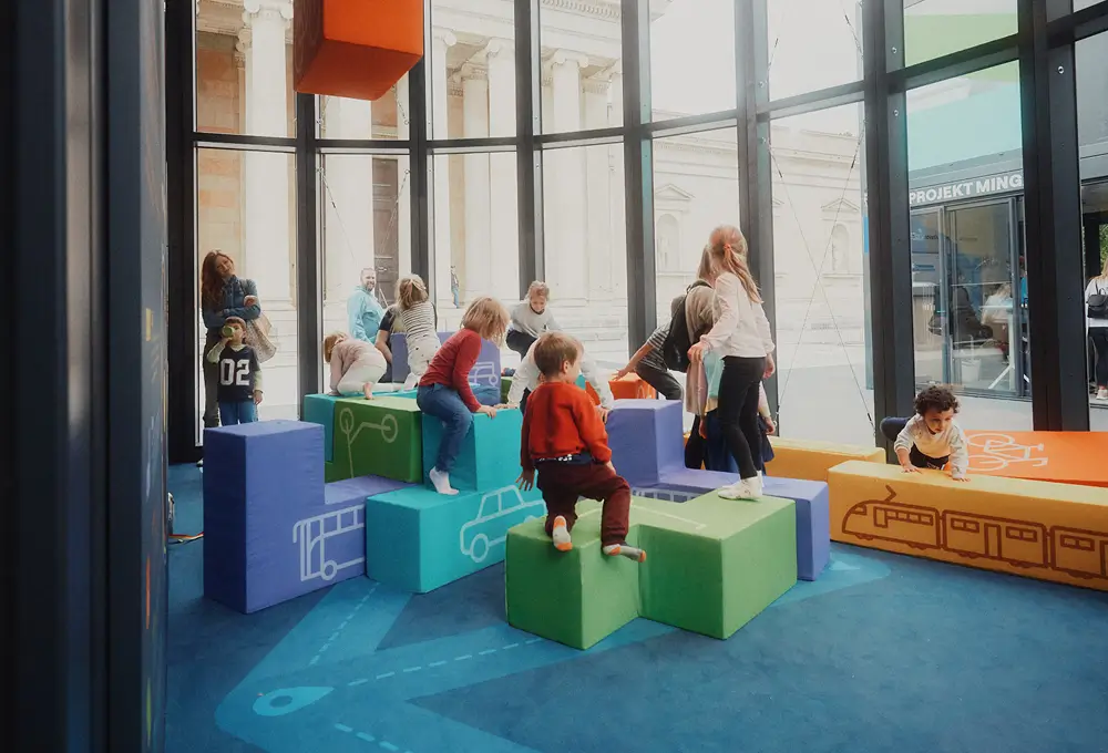 Kinder spielen auf bunten, würfelförmigen Kletterelementen mit Verkehrs- und Mobilitätsmotiven in einem hellen Raum.