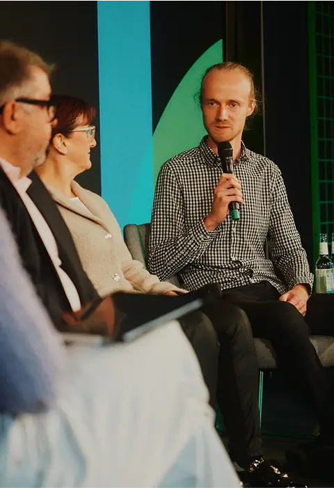 Mehrere Personen sitzen auf einem Podium, eine Person mit kariertem Hemd spricht in ein Mikrofon.