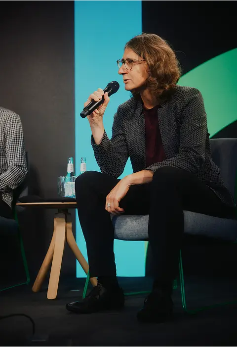 Eine Frau sitzt auf einem Podium und spricht in ein Mikrofon, neben ihr steht ein Glas Wasser.
