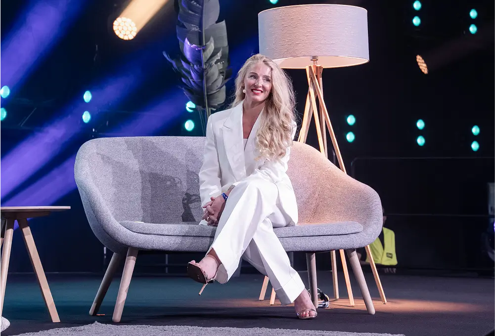 Eine Frau mit langen, blonden Haaren sitzt in einem weißen Hosenanzug auf einem Sofa auf einer Bühne mit Scheinwerfern und Stehlampe im Hintergrund.