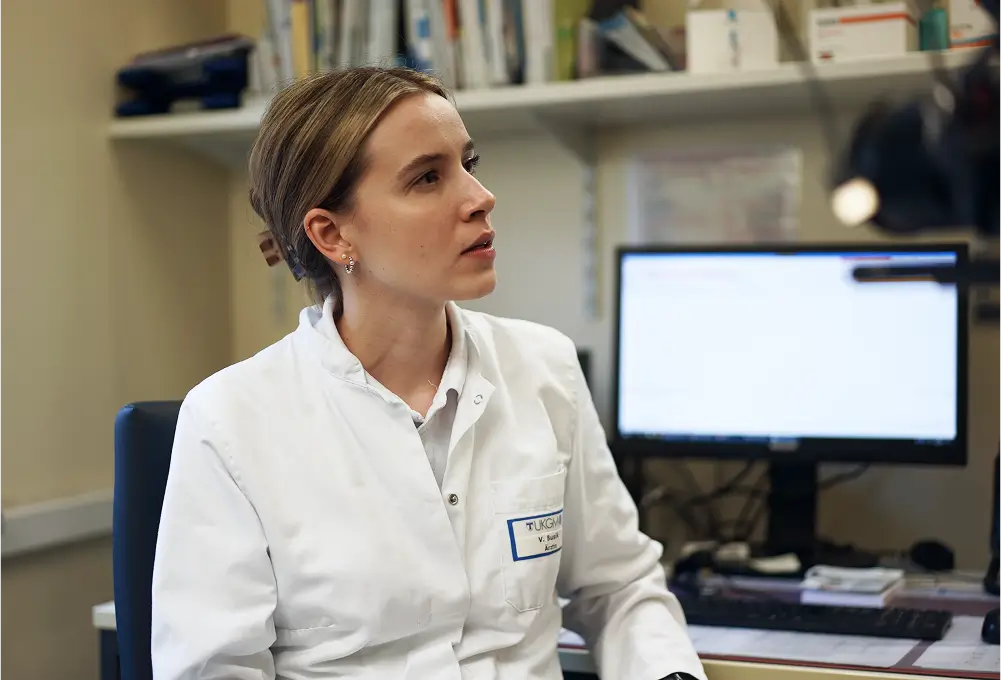 Eine Ärztin im weißen Kittel sitzt an einem Schreibtisch in einem Büro mit Computer und Bücherregal im Hintergrund. Sie trägt ein Namensschild und befindet sich in einer typischen Arztpraxis-Umgebung.