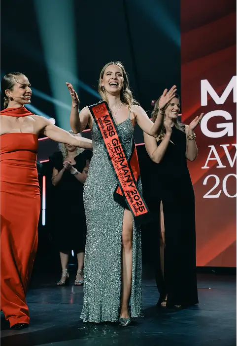 Eine Frau in einem glitzernden Abendkleid trägt eine Schärpe mit der Aufschrift „Miss Germany 2025“ auf einer Bühne bei einer Preisverleihung.
