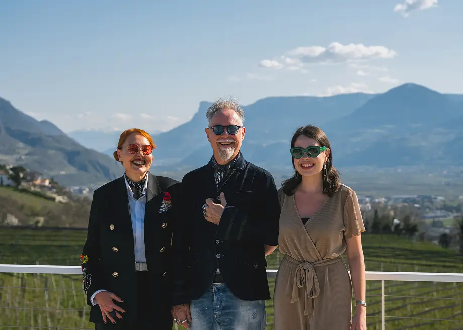 Drei lächelnde Personen mit Sonnenbrillen posieren auf Terrasse vor Südtiroler Bergpanorama.