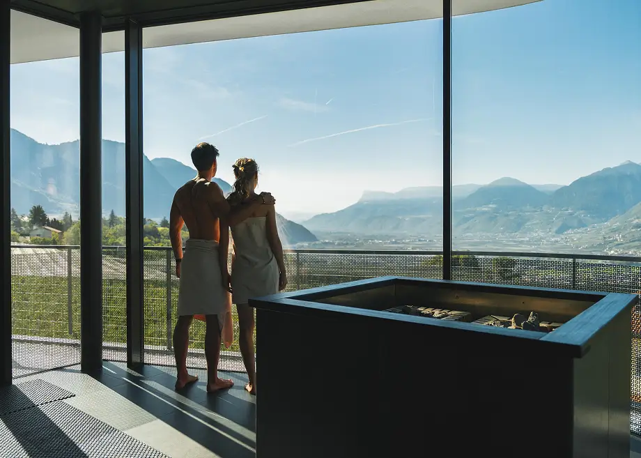 Ein Paar in Bademänteln blickt durch Panoramafenster auf Berglandschaft im Spa-Bereich.