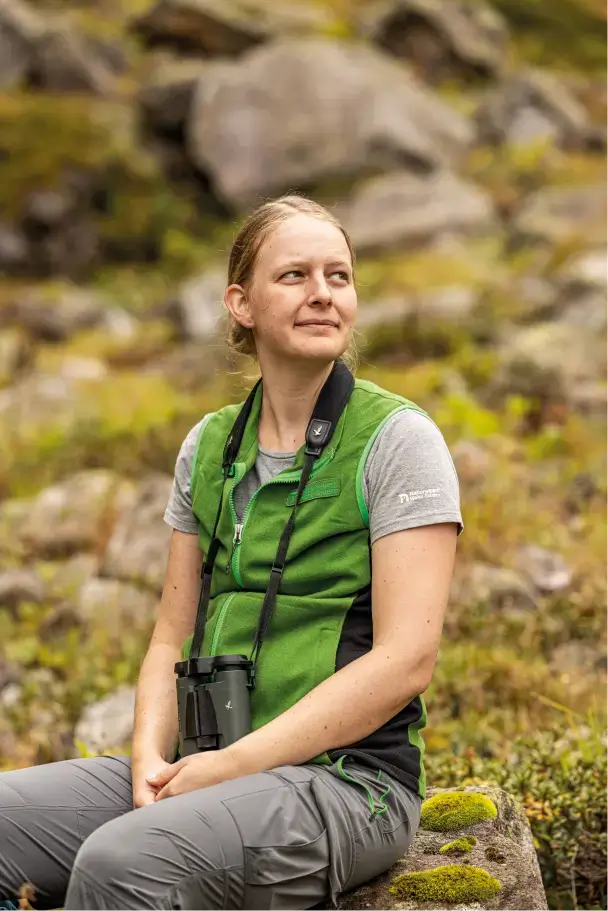 Sylvia Flucher sitzt auf einem Felsen in der Natur, trägt eine grüne Weste und hat ein Fernglas um den Hals.