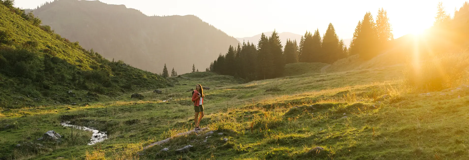 Eine Wanderin steht in einer sonnigen Berglandschaft auf einer grünen Wiese, umgeben von Hügeln und Bäumen. Warmes Abendlicht fällt über die Szene.