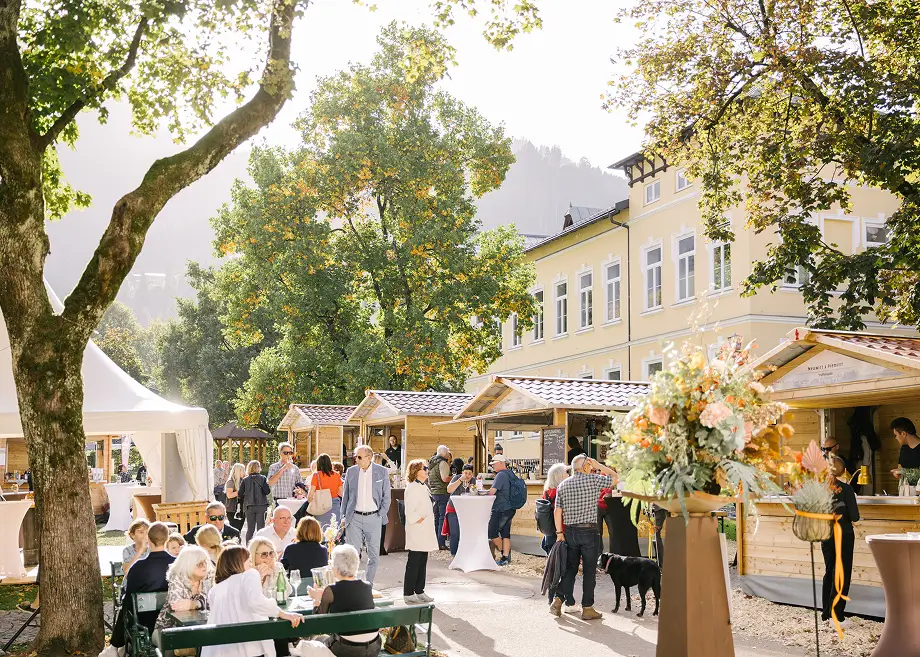 Menschen sitzen und stehen an Marktständen in einem sonnigen Platz mit Bäumen und hellem Gebäude im Hintergrund.