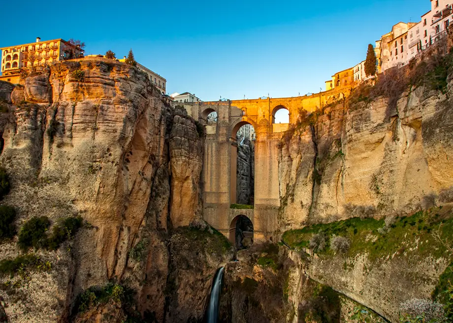Blick auf die Puente Nuevo, eine historische Bogenbrücke in Ronda, Spanien, die eine tiefe Felsschlucht mit einem kleinen Wasserfall überspannt. Auf beiden Seiten der Schlucht sind alte Gebäude unter blauem Himmel zu sehen.