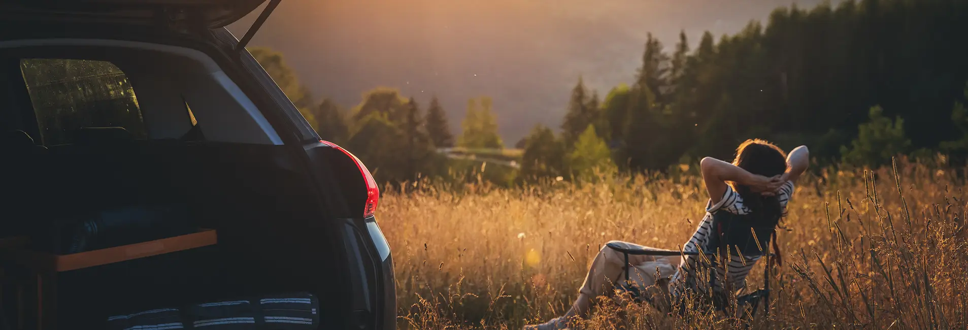 Eine Person sitzt entspannt in einem goldenen Getreidefeld neben einem parkenden SUV mit geöffneter Heckklappe. Im Hintergrund erstreckt sich eine Berglandschaft im Abendlicht.