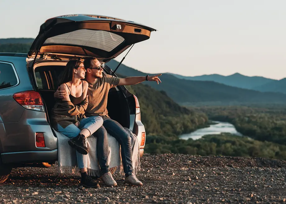 Ein Pärchen sitzt lächelnd in der geöffneten Heckklappe eines Autos auf einem Aussichtspunkt. Der Mann zeigt in die Ferne auf eine Flusslandschaft mit Bergen im Sonnenuntergang.