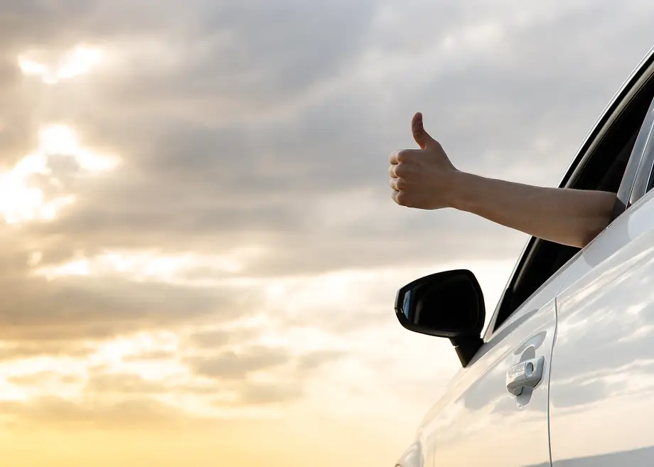 Eine Hand streckt sich aus dem geöffneten Fenster eines weißen Autos und zeigt einen Daumen hoch. Im Hintergrund ist ein bewölkter Himmel im warmen Abendlicht zu sehen.