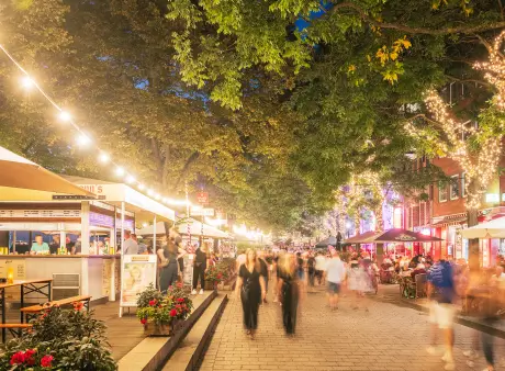 Belebte Promenade mit Marktständen, Restaurants und vielen Menschen unter beleuchteten Bäumen am Abend.