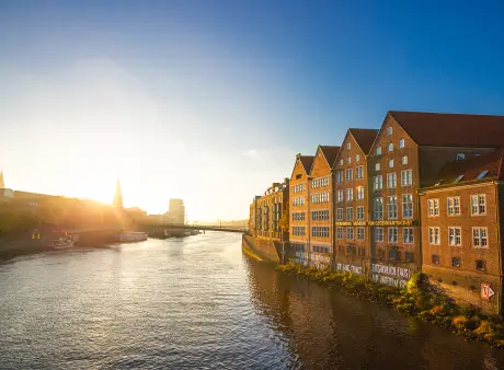 Blick über einen Fluss auf historische Häuserzeilen am Ufer und eine Brücke im warmen Abendlicht.