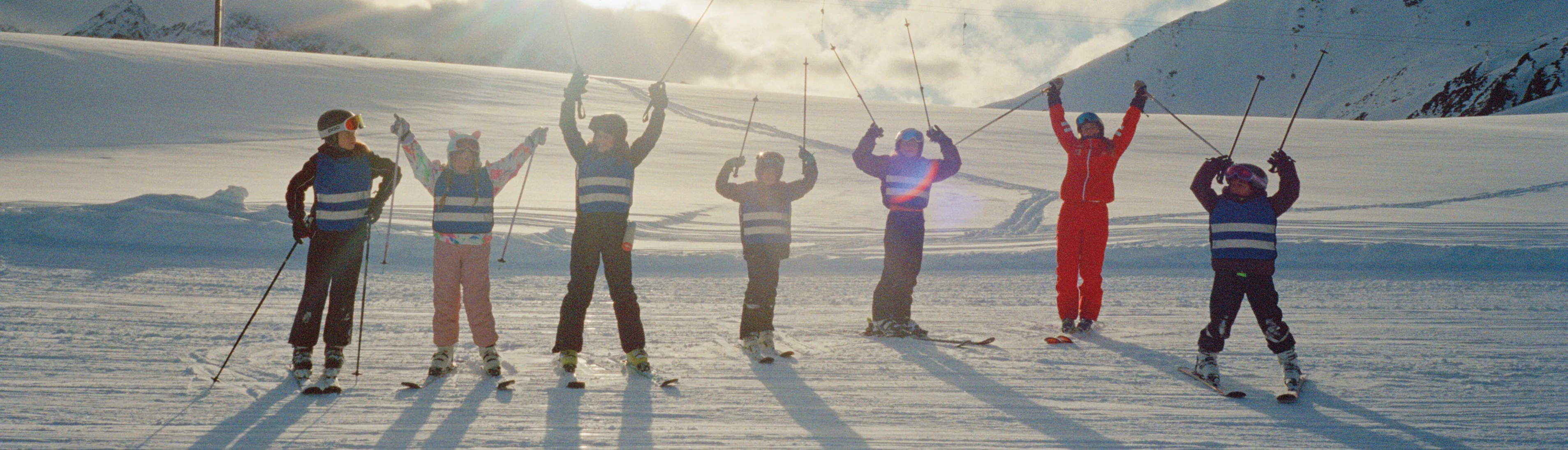 Sieben Skifahrerinnen und Skifahrer stehen auf einer verschneiten Piste und heben ihre Skistöcke oder Arme. Im Hintergrund liegen schneebedeckte Berge.