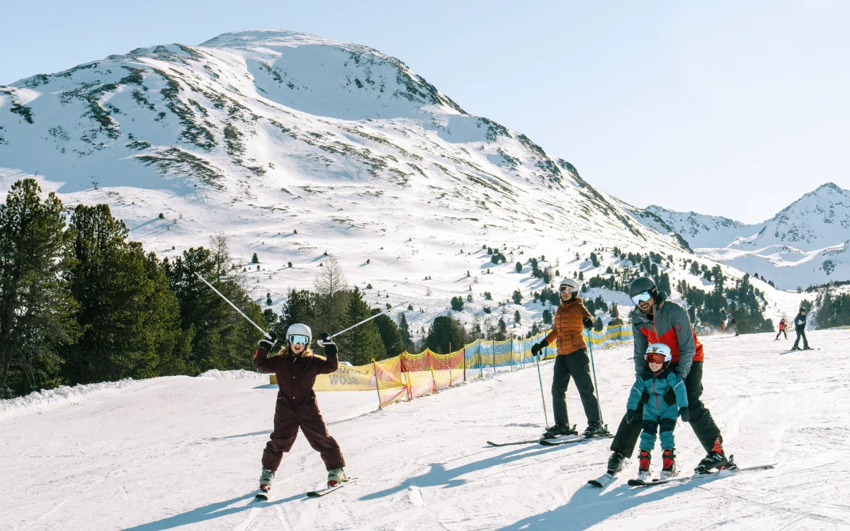 Vier Personen stehen auf einer Skipiste vor einem verschneiten Berg; eine Person im Vordergrund hebt die Skistöcke, während ein Erwachsener einem Kind beim Skifahren hilft. Weitere Skifahrer sind im Hintergrund zu sehen.
